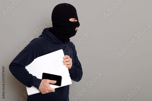 Side view of a criminal guy escaping with a stolen laptop, a woman's bag and a wallet, the robber is wearing a black hoodie, his face is disguised, copy space, isolated on gray background