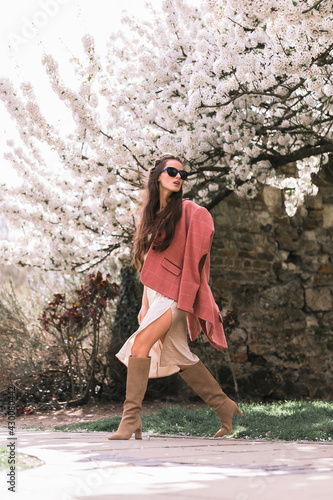 Beautiful young brunette girl, model, in a fashionable dress and a classic checkered pink jacket. Sunglasses. In motion. Looking away. Background of the alley of flowering trees.