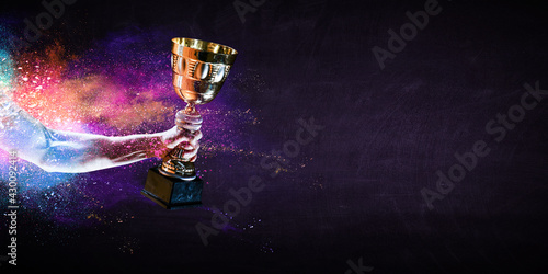 Fototapeta Hand holding up a gold trophy cup against dark background
