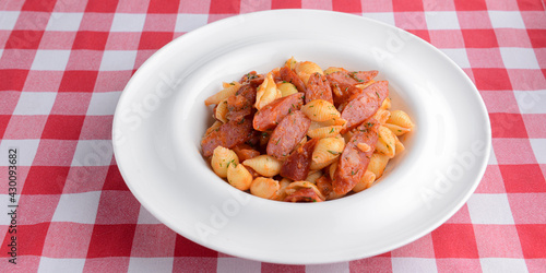 Plate of penne pasta with tomato sauce and meat sausages. Served in a white plate over red plaid table cloth.