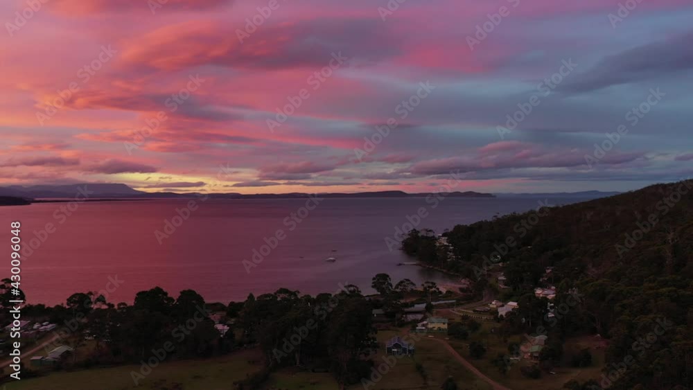 Slow aerial drone pan around purple stunning sunset island beach at dusk, purple and orange sky and mountain range with small shacks and forest. Adventure Bay, Bruny Island, Tasmania, Australia. Slow aerial drone pan around purple stunning sunset island beach at dusk, purple and orange sky and mountain range with small shacks and forest. Adventure Bay, Bruny Island, Tasmania, Australia.