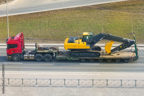 Heavy yellow excavator on transportation truck with long trailer platform on the highway in the city.