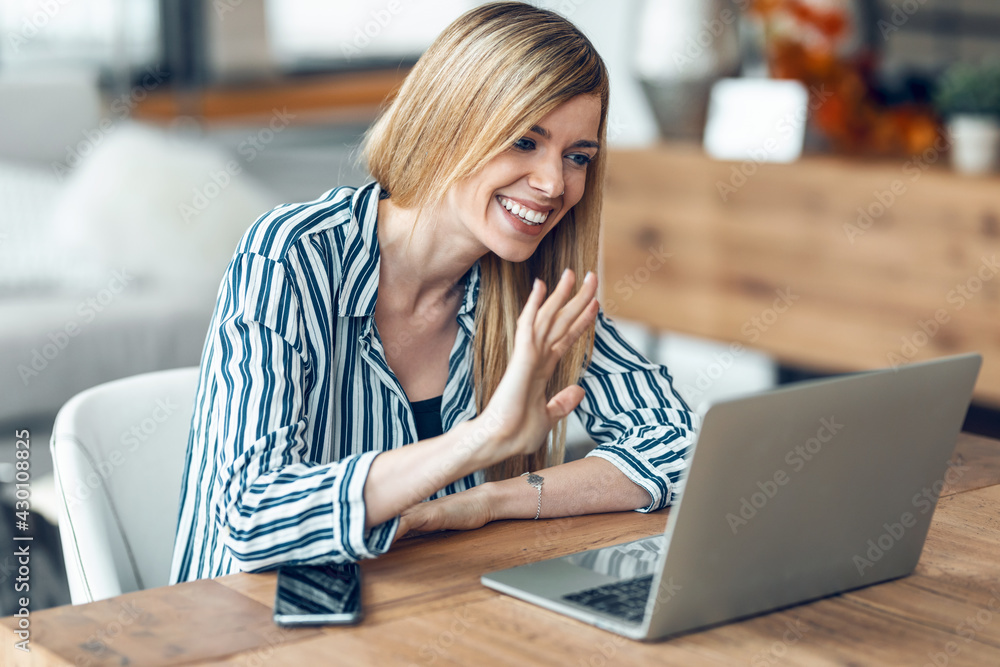 Fototapeta premium Smiling young woman waving hand while having videocall with laptop sitting in living room at home.