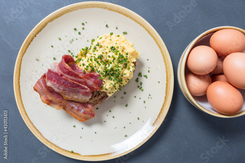Scrambled eggs with bacon and chives on sourdough toast on plate with bowl of eggs to one side. Top down shot.