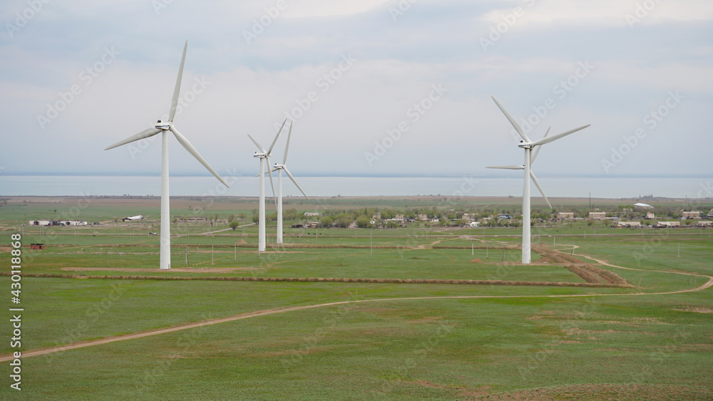 Windmills stand in the middle of green fields. Clean energy is generated. Cloudy weather, dark clouds. Green grass grows. High hills in the distance. Top view from drone. Industrial farm of Kazakhstan Windmills stand in the middle of green fields. Clean energy is generated. Cloudy weather, dark clouds. Green grass grows. High hills in the distance. Top view from drone. Industrial farm of Kazakhstan