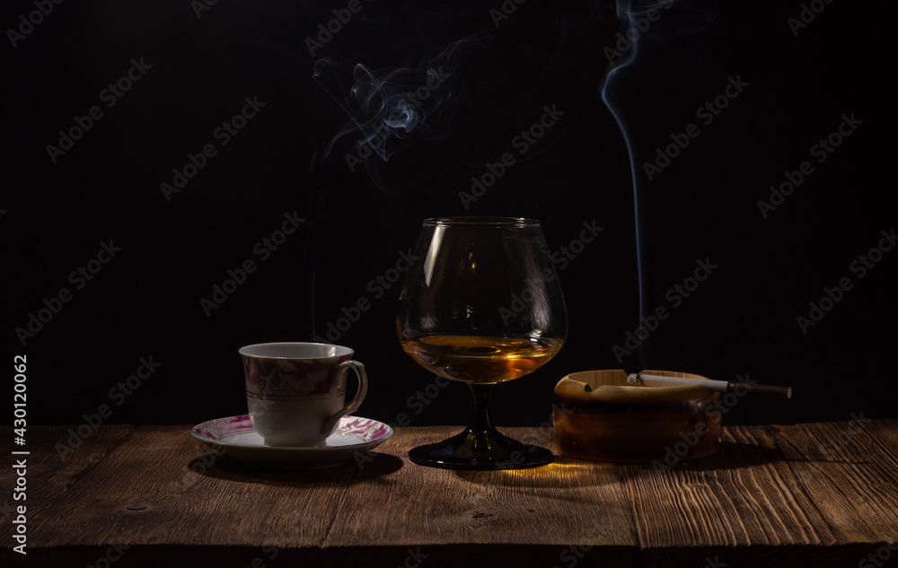 Background image of a wooden bar in a pub with a glass of cognac and a smoking cigarette in an ashtray and a cup of coffee on a dark background with a bokeh effect Background image of a wooden bar in a pub with a glass of cognac and a smoking cigarette in an ashtray and a cup of coffee on a dark background with a bokeh effect