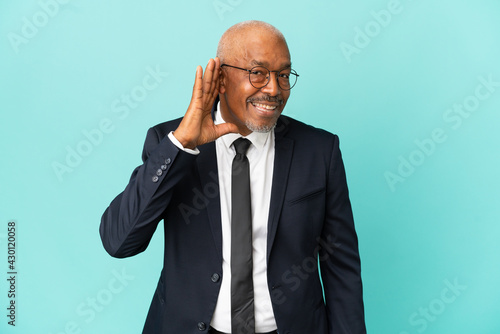 Business senior man isolated on blue background listening to something by putting hand on the ear