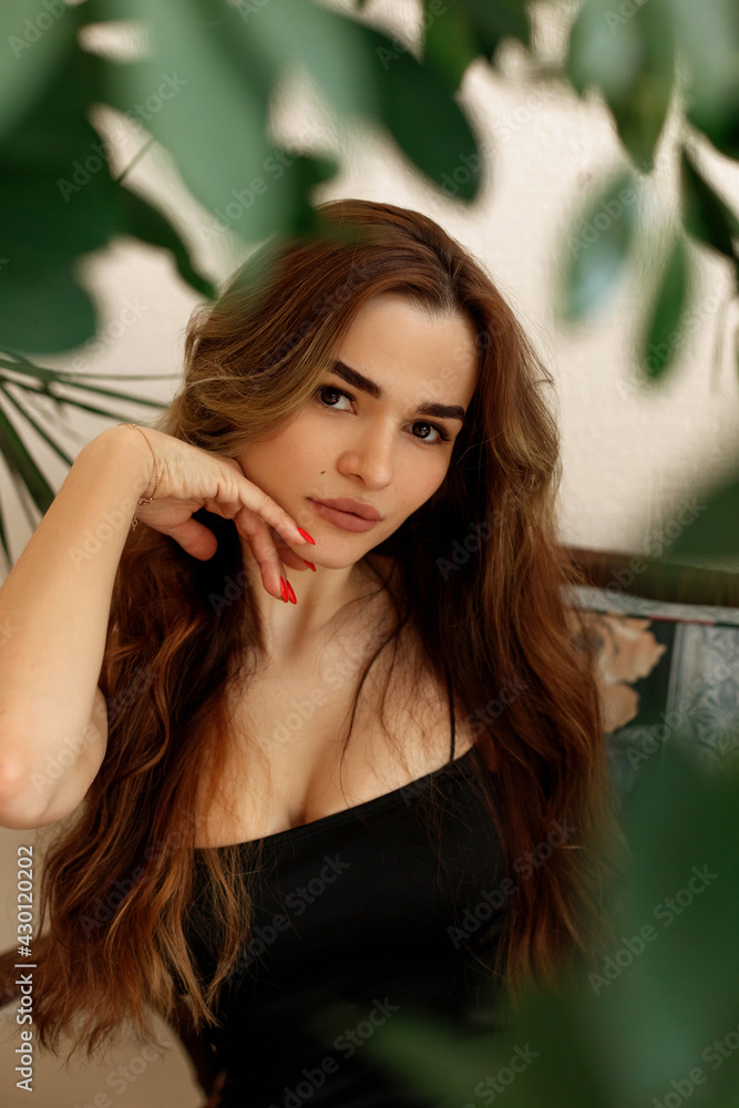 Fototapeta premium Beautiful women in greenery looking at the camera, sitting on the chair on terrace at home. Portrait of a girl with long hair brown-haired, tropical ethno boho style interior
