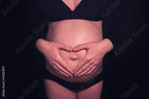 Pregnant woman belly with heart-shaped hands, front view on black background