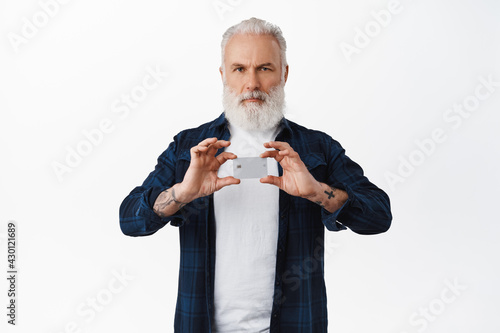 Serious senior man with long white beard and tattoos, shows credit card and frowns self-assured, stands against white background © Cookie Studio