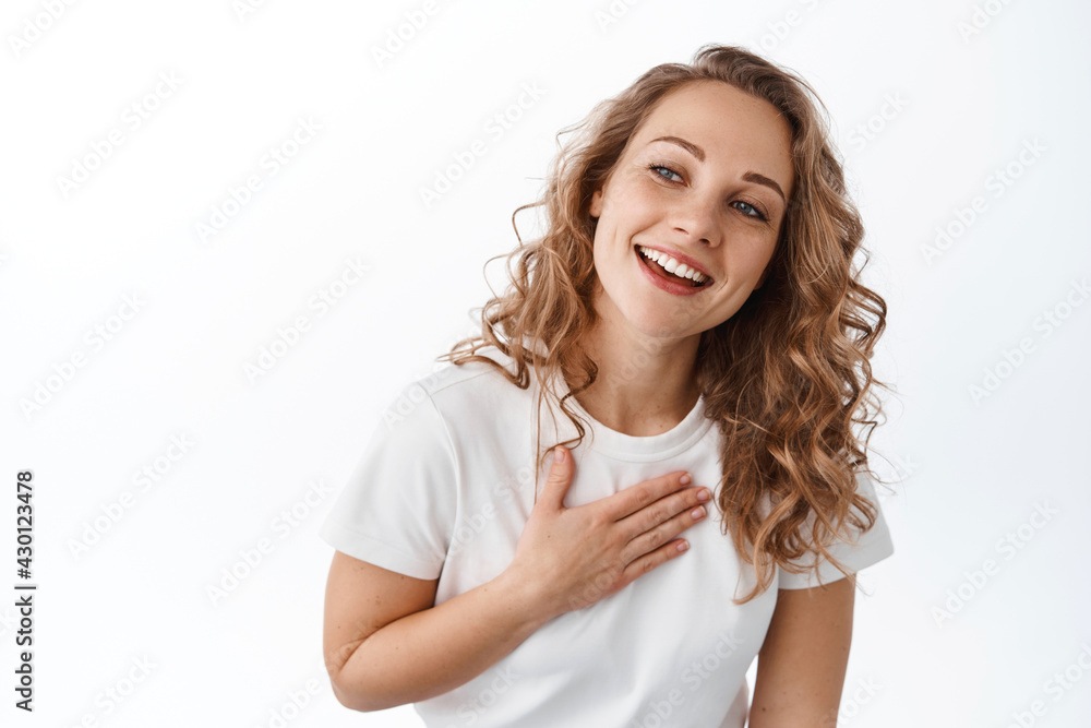 Grateful blond girl smiling, holding hand on heart and looking aside at promotional text with thankful and pleased face, standing over white background Grateful blond girl smiling, holding hand on heart and looking aside at promotional text with thankful and pleased face, standing over white background