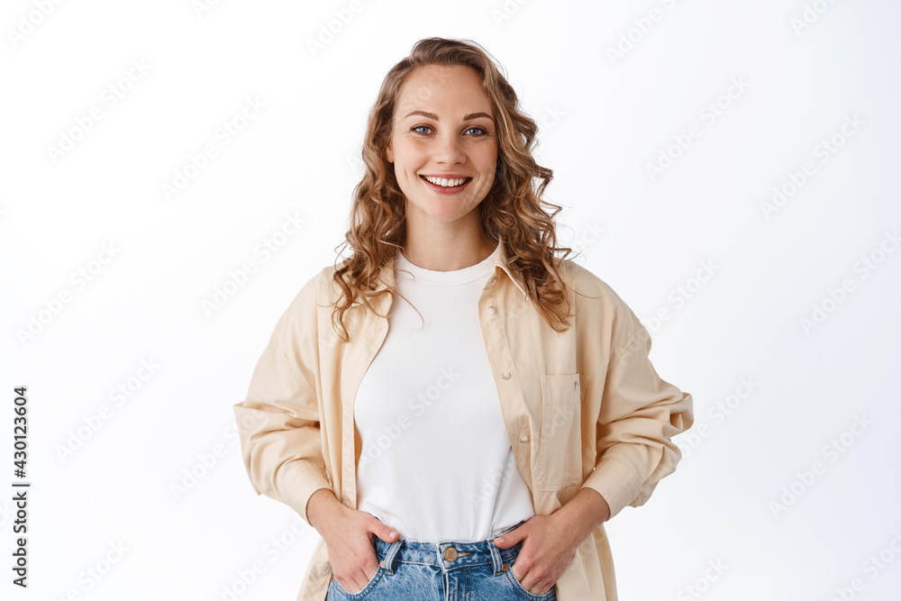 Obraz premium Stylish young modern woman with blond hairdo, holding hands in jeans pockets and smiling carefree, looking confident and determined, white background