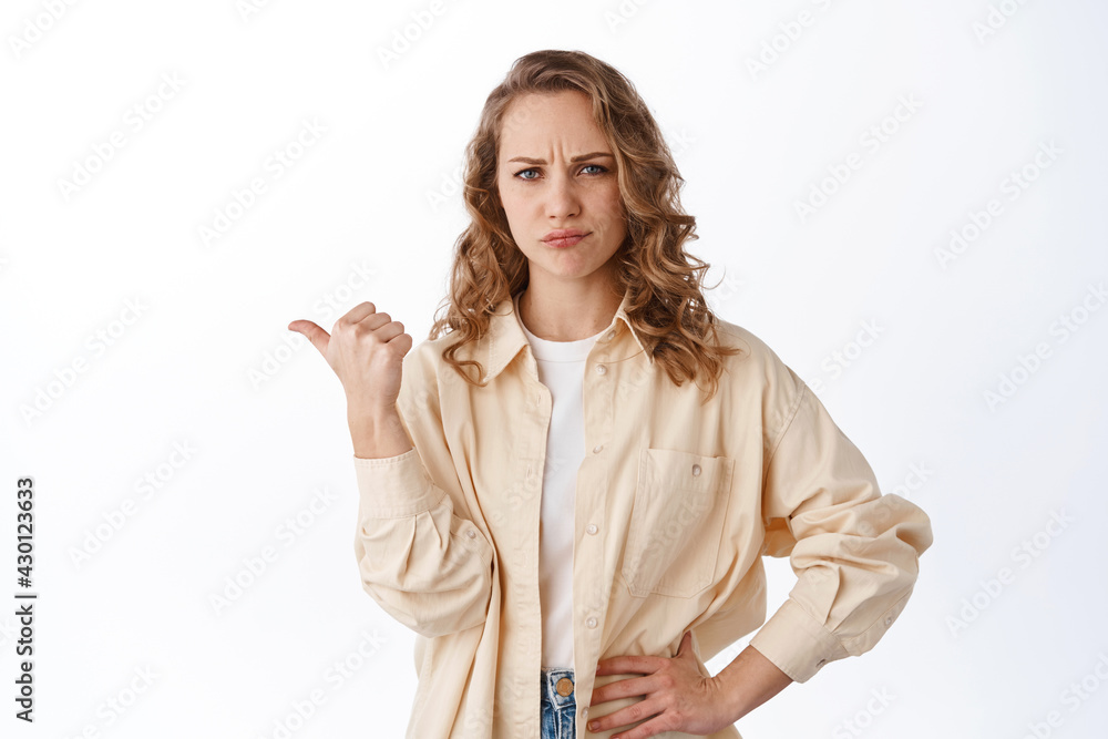 Doubtful blond woman grimacing, pointing finger left at copyspace, being uncertain or displeased, look skeptical, standing over white background Doubtful blond woman grimacing, pointing finger left at copyspace, being uncertain or displeased, look skeptical, standing over white background