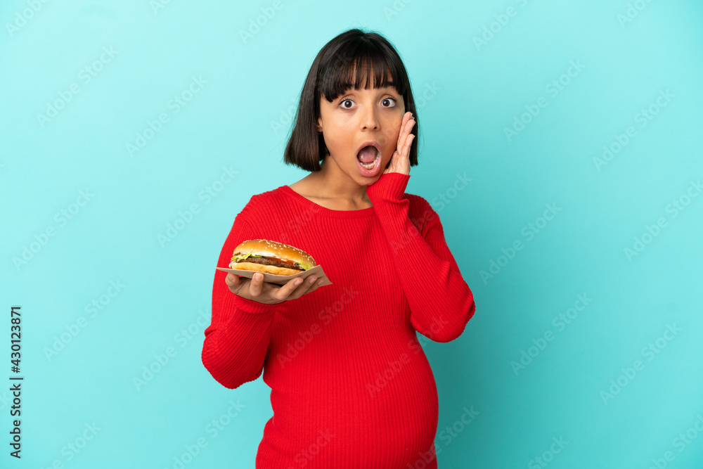 Young pregnant woman holding a burger over isolated background with surprise and shocked facial expression Young pregnant woman holding a burger over isolated background with surprise and shocked facial expression