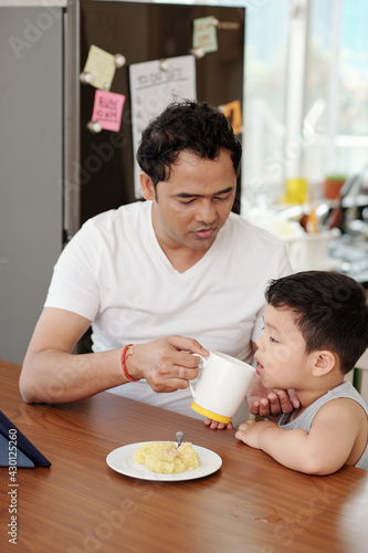 Father giving his little son a cup of water when they are eating breakfast and watching animated film on tablet computer