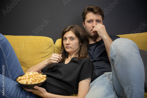 Couple bored watching tv movie hugging lying on couch with popcorn in hands, YouTube channel, online broadcast. Evening entertainment for young family. Unhealthy habits lead to weight gain