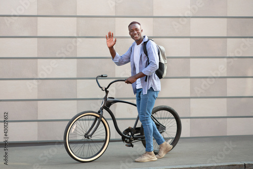 Positive millennial guy with modern bike waving near brick wall outdoors