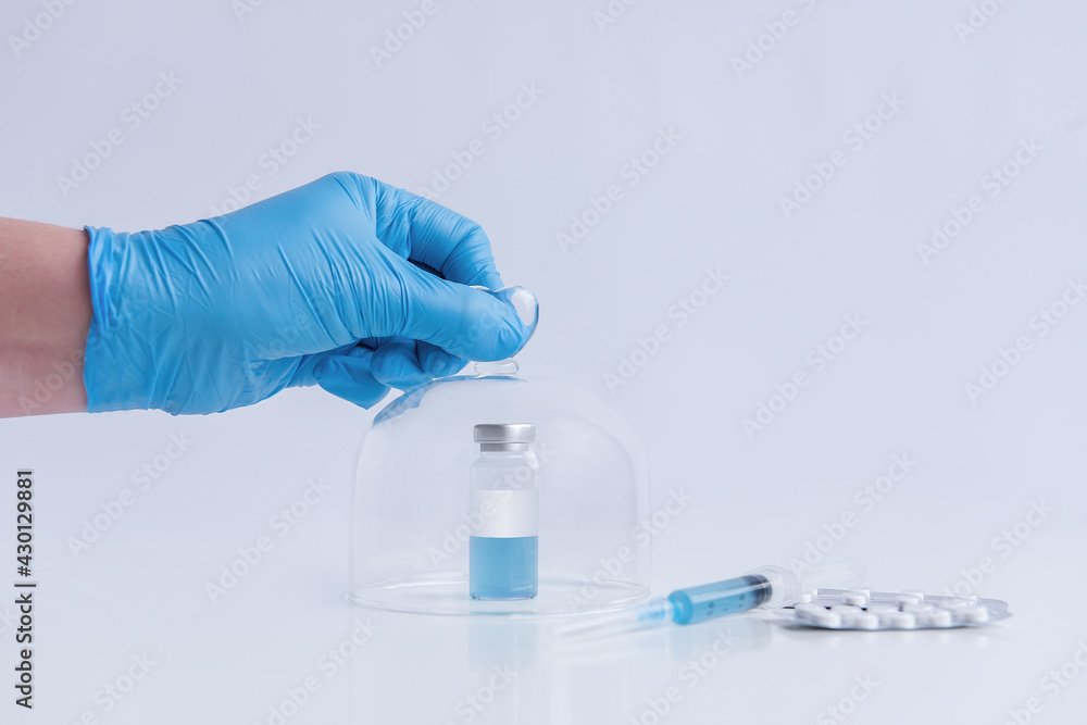 On a white isolated background stands the vaccine with a syringe, pills. The doctor's hand in a blue medical glove opens the programmed protective dome. Cure against viruses, treatment of people On a white isolated background stands the vaccine with a syringe, pills. The doctor's hand in a blue medical glove opens the programmed protective dome. Cure against viruses, treatment of people