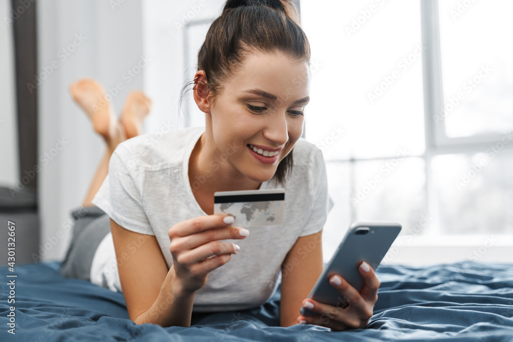 Smiling woman holding credit card and using cellphone while lying on bed Smiling woman holding credit card and using cellphone while lying on bed