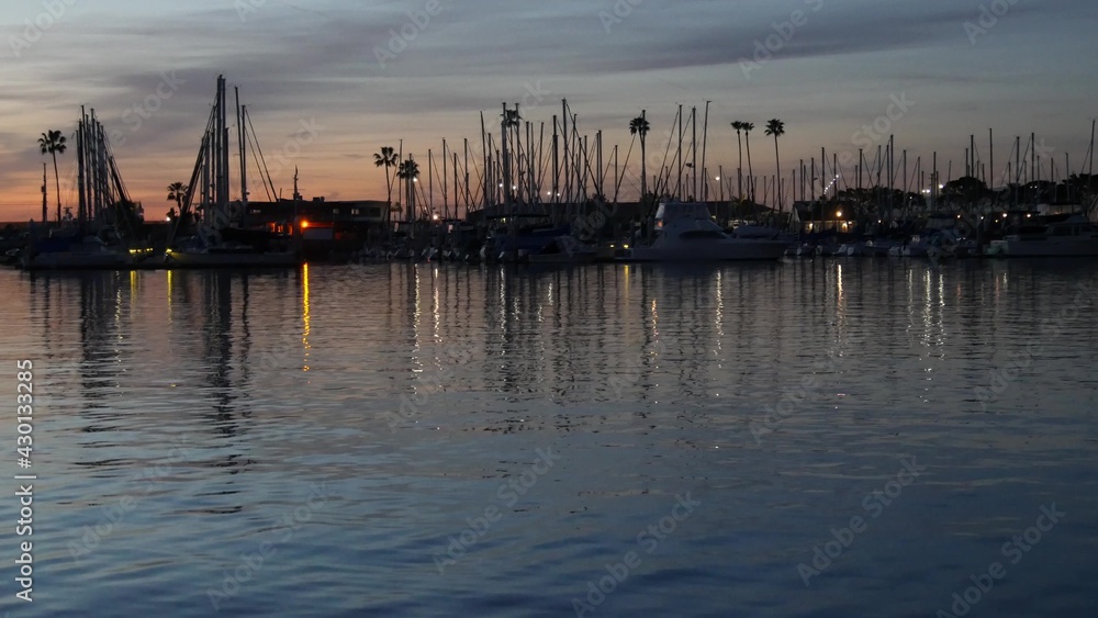 Luxury yachts sailboats floating, marina harbour quay. Sail boat masts, nautical vessels in port. Harbor fisherman village in Oceanside, California USA. Evening dusk, twilight lights and palm trees.
