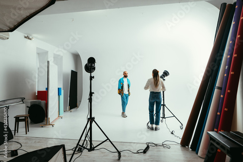 Woman photographer with a camera in his hands in a photo studio makes a photo shoot for a handsome bearded man on a white background, uses flashes.