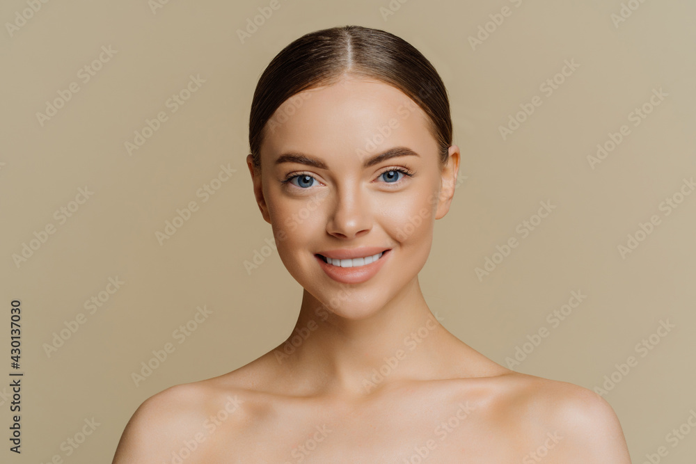 Beauty portrait of charming European woman with beautiful face healthy complexion well cared body smiles gently stands with bare shoulders indoor against beige background has clean fresh skin Beauty portrait of charming European woman with beautiful face healthy complexion well cared body smiles gently stands with bare shoulders indoor against beige background has clean fresh skin