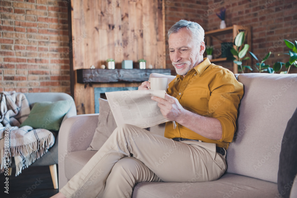 Photo of positive aged man sit on couch holding coffee reading interested newspaper enjoy home indoors