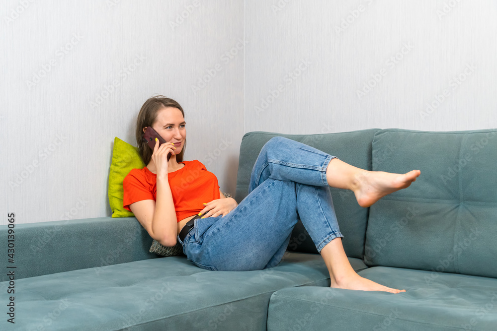 Young beautiful dark-haired woman in blue jeans and an orange shirt is lying on the sofa and talking on the phone Young beautiful dark-haired woman in blue jeans and an orange shirt is lying on the sofa and talking on the phone