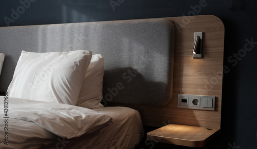 Close-up fragment of bedroom with empty bedside table, reading lamp and a USB socket in modern interior​ design home or hotel. Soft pillow and blanket, stylish comfortable furniture. Sun shadows.