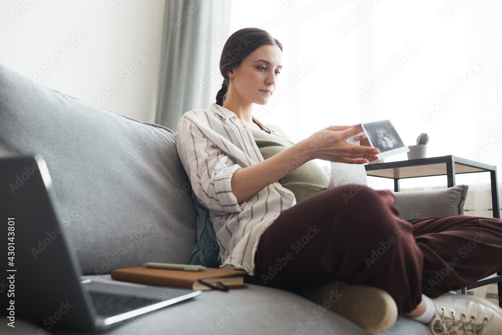 Young pregnant woman sitting on a couch with laptop and looking at x-ray image of her baby Young pregnant woman sitting on a couch with laptop and looking at x-ray image of her baby