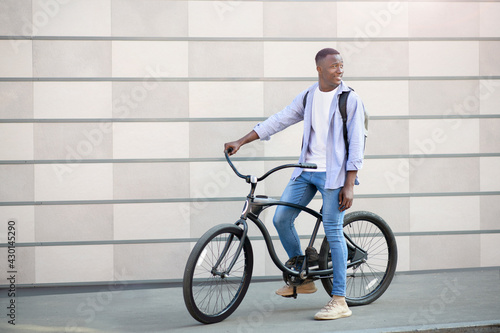 Urban adventure. Happy black man with backpack and bike standing near brick wall outdoors and looking aside, copy space