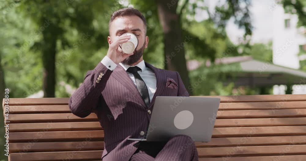 Confident mature businessman wearing suit working on laptop remotely, sitting on bench in park and drinking coffee Confident mature businessman wearing suit working on laptop remotely, sitting on bench in park and drinking coffee