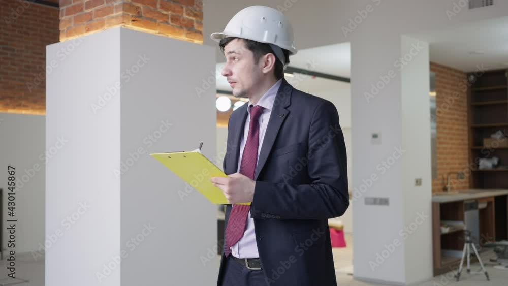Focused man in hard hat and suit filling in documents standing in new apartment under construction. Portrait of professional concentrated foreman checking repair works indoors. Real estate profession Focused man in hard hat and suit filling in documents standing in new apartment under construction. Portrait of professional concentrated foreman checking repair works indoors. Real estate profession
