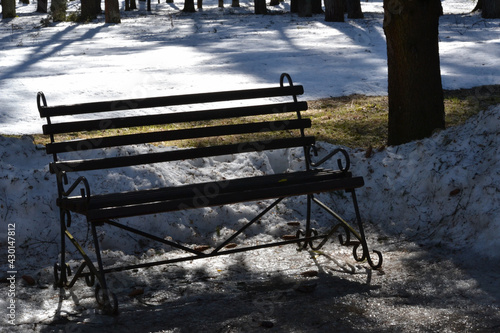 In the city park in spring. A beautiful bench in the shade of a park alley in spring against the backdrop of melting snow.