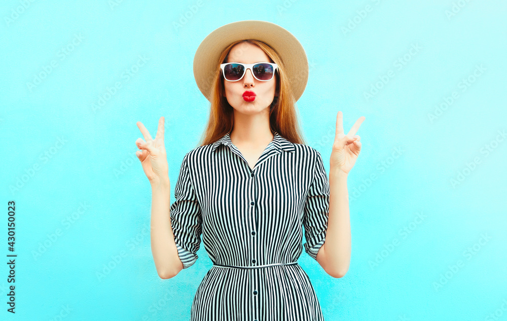 Portrait of beautiful young woman blowing her red lips wearing a striped dress, summer straw hat on a blue background Portrait of beautiful young woman blowing her red lips wearing a striped dress, summer straw hat on a blue background