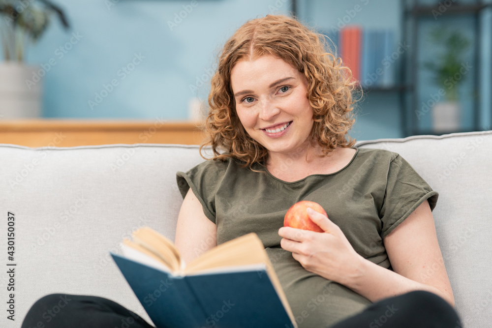 Smiling pregnant woman sitting on sofa reading book and holding apple in one hand. Smiling pregnant woman sitting on sofa reading book and holding apple in one hand.