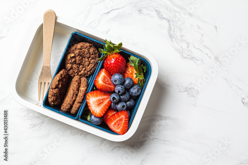 Fototapeta Naklejka Na Ścianę i Meble -  Lunch box with fruits and cookies on white marble background, top view. Takeaway healthy snack container.
