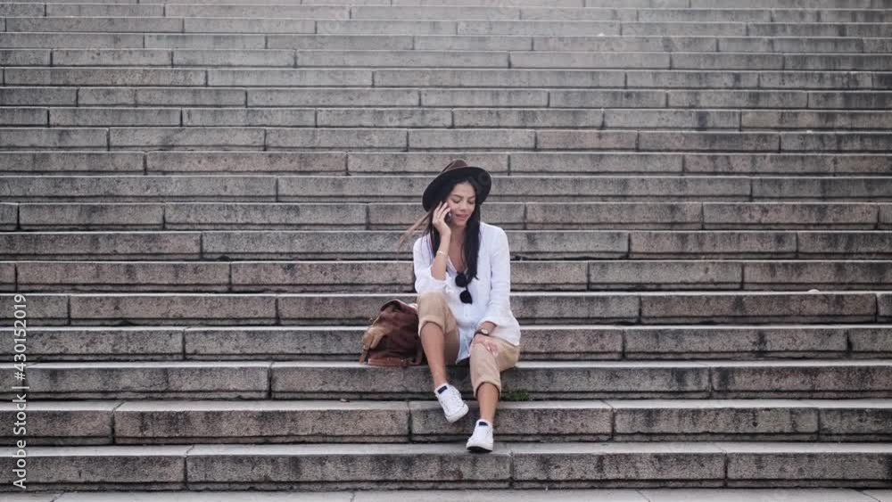 A beautiful young woman wearing a hat is talking on her mobile phone sitting on the stair outside in summer A beautiful young woman wearing a hat is talking on her mobile phone sitting on the stair outside in summer