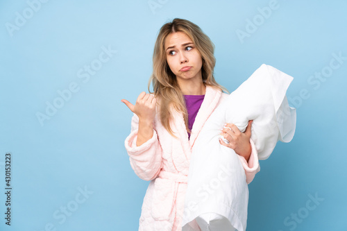 Teenager Russian girl in pajamas isolated on blue background unhappy and pointing to the side