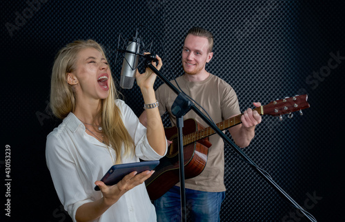 Young female singing with tablet computer in her hand. While the man playing an acoustic guitar in front of black soundproofing walls. Musicians producing music in professional recording studio.