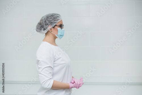 female doctor wearing glasses, a white medical suit, a cap, a medical mask, and disposable gloves stands sideways in the office