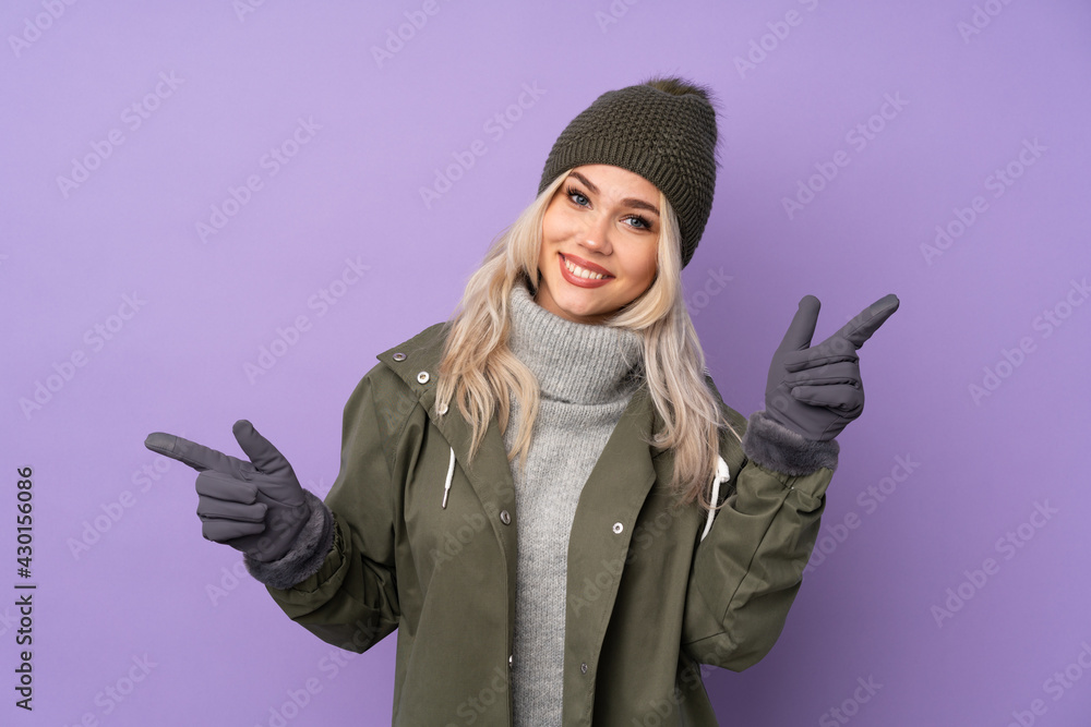 Teenager blonde girl with winter hat over isolated purple background pointing finger to the laterals and happy Teenager blonde girl with winter hat over isolated purple background pointing finger to the laterals and happy