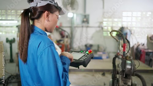 Robotic arm machine was controlled  by engineer woman in the blue uniform standing in the factory. Wearing safety helmet and glasses. Industrial, technology and innovation lifestyle for ai concept