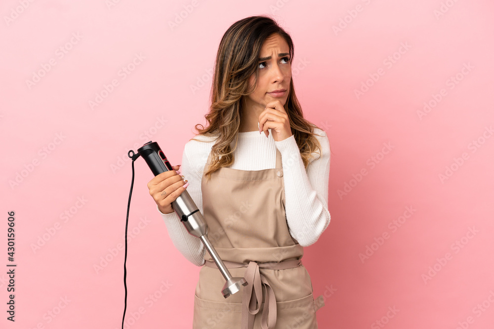 Young woman using hand blender over isolated pink background and looking up