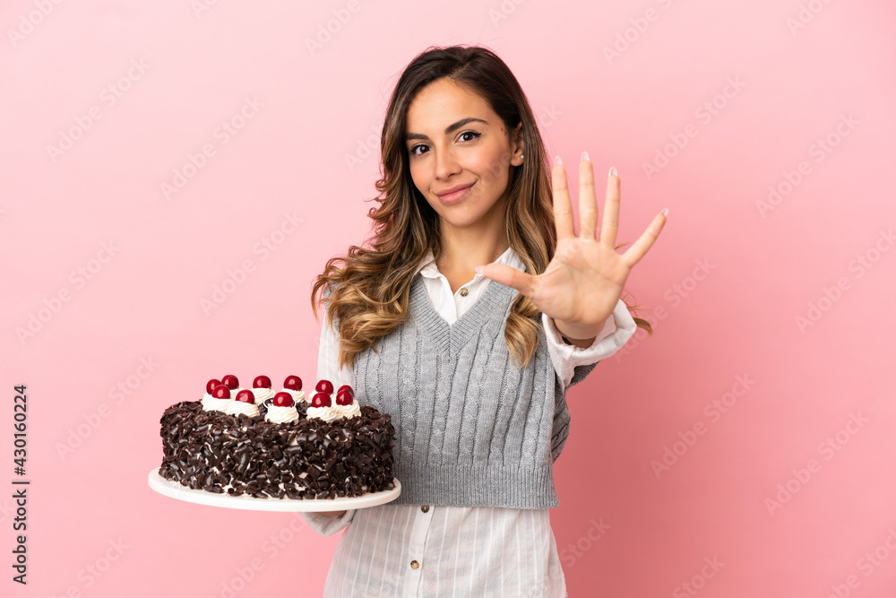 Young woman holding birthday cake over isolated pink background counting five with fingers Young woman holding birthday cake over isolated pink background counting five with fingers