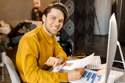 Stylish happy young businessman sitting at the desk, working from home office, learning financial graphs. Successful male freelancer doing some paperwork, looking at the camera, smiling