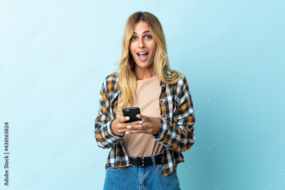 Young blonde Uruguayan isolated on blue background surprised and sending a message Young blonde Uruguayan isolated on blue background surprised and sending a message
