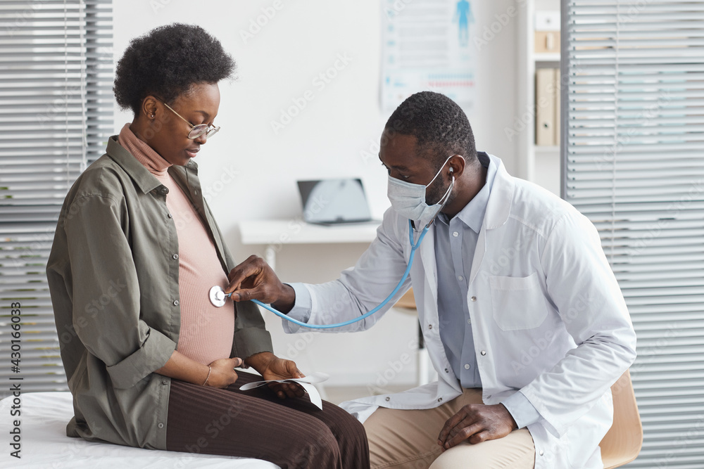 African doctor in white coat examining the belly of pregnant woman with stethoscope during visit at hospital - obrazy, fototapety, plakaty 