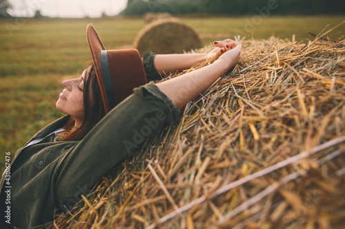 Fototapeta Beautiful stylish woman in hat relaxing on haystack in summer evening field