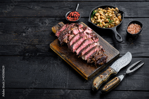 Juicy medium beef Rib Eye steak slices, on wooden serving board, with white beans and rosemary in cast iron pan, with meat knife and fork, on black wooden table background, with copy space for text